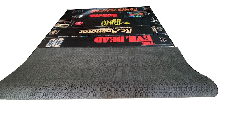 May include: A black and gray VHS tape rug with the titles "The Evil Dead", "Re-Animator", "The Thing", "Madman", and "Pumpkinhead" printed on the top.