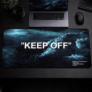 Puede incluir: Alfombrilla de escritorio rectangular negra con un diseño de nubes azules y blancas y las palabras "KEEP OFF" en blanco. Un portátil, un ratón y un teclado son visibles en el fondo.