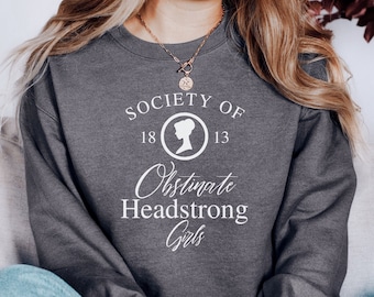 May include: A gray sweatshirt with the text "Society of Obstinate Headstrong Girls" and the numbers "18" and "13" on a white circle with a silhouette of a woman's head inside.