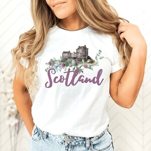 May include: A white t-shirt with a watercolor illustration of a castle and flowers. The text "Scotland" is written in purple below the illustration.