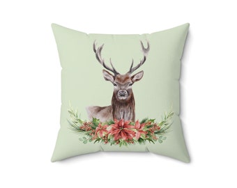 Puede incluir: Una almohada verde con una ilustración de acuarela de un ciervo con cuernos rodeado de una corona de flores de poinsettia rojas y hojas verdes.