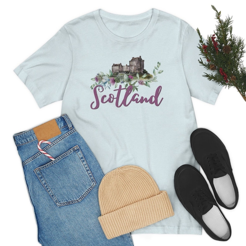 May include: Light blue t-shirt with a watercolor illustration of a castle and flowers. The text "Scotland" is written in purple cursive lettering.