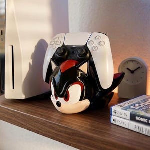 May include: A black, red, and white Shadow the Hedgehog-themed controller holder with a white PlayStation 5 controller resting on top. Two video game cases and a small clock are also visible on the wooden shelf.