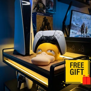 May include: A white and black PlayStation 5 console and a matching wireless controller resting on a yellow Psyduck-shaped stand. The controller is positioned on top of the yellow character head. A yellow square graphic with the text FREE GIFT is visible.