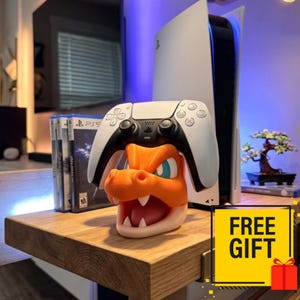 May include: An orange and cream-colored dragon head controller stand holding a white and black wireless game controller. The stand features a wide-open mouth with visible teeth and blue eyes, positioned in front of a white gaming console and game cases.