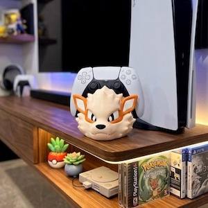 May include: A cream-colored Arcanine figure with orange accents, positioned in front of a white PlayStation 5 controller. The figure is on a dark wood shelf, alongside a PS5 console and video games. The shelf also features small potted plants and a retro game console.