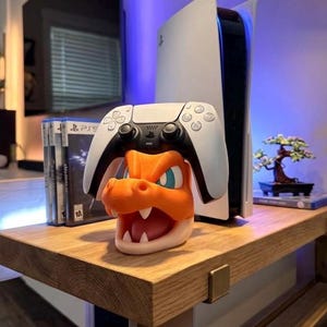 May include: A white PlayStation 5 controller rests on an orange and white dragon-themed console stand. A white PlayStation 5 console and game cases are visible in the background. The stand is on a wooden table.
