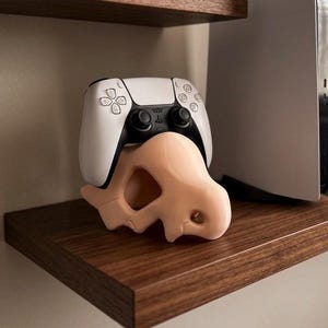 May include: A white and black video game controller rests on a light pink, skull-shaped stand. The controller is on a dark brown wooden shelf, with another shelf above it.