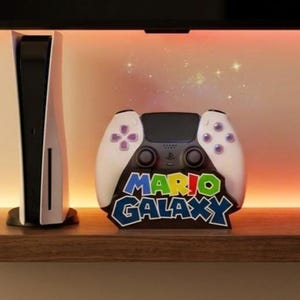 May include: A white and black gaming console and a controller-shaped stand. The stand features the words "MARIO GALAXY" in a colorful, outlined font. The controller has white grips and purple button accents.