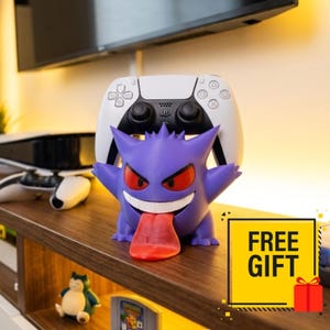 May include: A purple Gengar character figure serving as a controller stand, holding a white and black wireless game controller. The figure features red eyes, a wide white grin, and a long red tongue extending downward from its mouth.