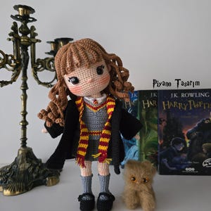 Puede incluir: Muñeca de ganchillo hecha a mano con cabello rizado castaño, vestida con un vestido gris, una túnica negra y una bufanda roja y dorada. Un pequeño gato de ganchillo marrón y dos libros de "Harry Potter" también están en la imagen. El texto "Piyano Tasarim" es visible.