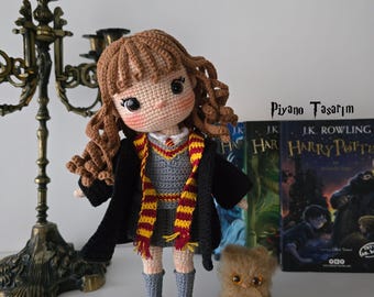 May include: A handmade crochet doll with brown curly hair, wearing a gray dress, black robe, and a red and gold scarf. A small, brown, fuzzy cat doll sits nearby. The doll is next to a stack of Harry Potter books.