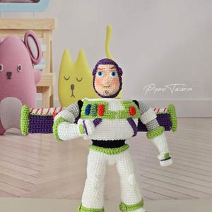 Op de afbeelding: Een gehaakte Buzz Lightyear-pop met een wit, groen en paars ruimtepak, een paarse kap, blauwe ogen en rode, blauwe en groene knoppen op de borst. Het figuur heeft uitgestrekte vleugels en staat rechtop, volledig gemaakt van garen.