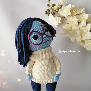 May include: A blue crocheted doll with long blue hair, wearing pink glasses, a white sweater, and blue pants. The doll is standing on a wooden surface with white flowers in the background. The text '@piyanotasarim' is visible in the bottom right corner of the image.