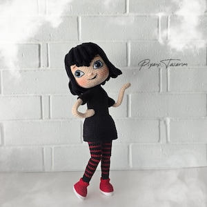 May include: A crocheted doll with black hair, a black dress, red and black striped leggings, and red shoes. The doll is standing on a white surface with a white brick wall in the background.