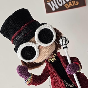 Puede incluir: Muñeca de ganchillo de un personaje con sombrero de copa negro con una raya roja, gafas grandes con montura blanca y abrigo rojo. La muñeca sostiene un bastón y lleva un collar dorado. Un letrero que dice "Wonka Bar" está en el fondo.