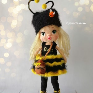 May include: A crocheted doll dressed as a bee with a black and yellow striped dress, black tights, yellow boots, and a honey pot purse. The doll has blonde hair, a black hat with a bee antenna, and a bee charm on the hat. The doll is standing on a wooden surface. The text "PIANO TASARIM" is visible in the upper right corner of the image.