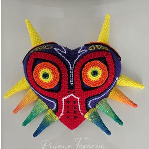 Op de afbeelding: Een gehaakt hartvormig masker in levendige kleuren. Het masker heeft een rode basis met oranje en gele concentrische cirkels voor de ogen, omlijnd in zwart. Het masker heeft gele en regenboogkleurige spikes. De tekst "Piyano Tasarim" staat onderaan.