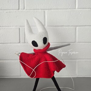 Pode incluir: Figura de croch&ecirc; de um personagem com cabe&ccedil;a branca, olhos pretos e capa vermelha. A figura tem uma espada cinza e pernas pretas. O texto "Piyano Tasarim" est&aacute; escrito em it&aacute;lico &agrave; direita.