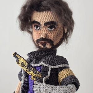 May include: Crocheted doll depicting a man with dark hair and a beard. The doll wears a black and gray outfit with gold accents, a purple shirt, and a sword. The doll's face is tan with dark eyes.