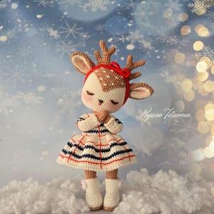 May include: A crocheted deer doll wearing a red bow and a plaid dress with white, blue, and red stripes. The doll is standing on a white fluffy surface with a blue and white snowflake background.