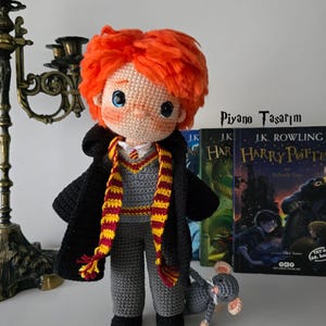 May include: A handmade crochet doll with orange hair, wearing a black robe, gray sweater, and scarf with red and yellow stripes. A small gray crochet mouse sits at the doll's feet. The doll is positioned in front of two "Harry Potter" books.