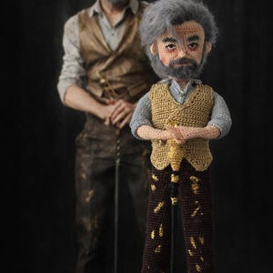 Può includere: Bambola all'uncinetto di un uomo con capelli grigi e barba, che indossa un gilet e tiene un bastone. La bambola è marrone, beige e grigia. Un uomo con un abito simile è in piedi sullo sfondo. La bambola è alta circa 30 cm.