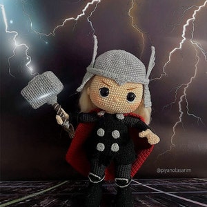Può includere: Un giocattolo di peluche a forma di Thor, un supereroe dei fumetti Marvel, che tiene un martello. La figura è vestita con un costume nero e grigio con un mantello rosso e un elmo grigio. Lo sfondo è grigio scuro con fulmini bianchi.