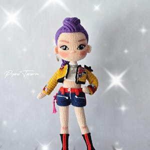 Könnte beinhalten: Eine handgefertigte Häkelpuppe mit violettem Haar in einem hohen Dutt, bekleidet mit einer gelben kurzen Jacke, einem weißen Oberteil, dunkelblauen Shorts mit rotem Saum und schwarzen Stiefeln mit roten vertikalen Streifen.