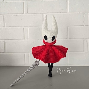 Op de afbeelding: Handgemaakte gehaakte pop met een witte kop, twee gebogen hoorns, zwarte ogen en een rode cape. De pop heeft zwarte benen en een grijze haaknaald. De tekst "Piyano Tasarim" is zichtbaar.