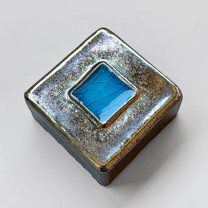 Puede incluir: Un azulejo de cerámica cuadrado con un esmalte metálico iridiscente. El azulejo tiene un cuadrado más pequeño insertado en el centro, que está esmaltado en un azul turquesa vibrante.