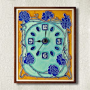 Op de afbeelding: Een blauwe en gele keramische klok met een bloemenmotief. De wijzerplaat van de klok is lichtblauw met witte cijfers en zwarte wijzers. De klok is ingelijst in een donkerbruin houten frame.