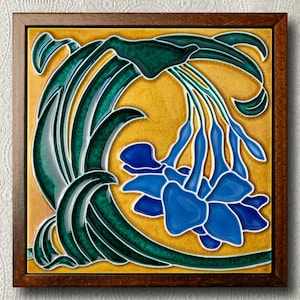 Può includere: Una piastrella di ceramica con un motivo floreale verde e blu. La piastrella è incorniciata da una cornice in legno scuro. Il motivo floreale presenta un fiore blu con foglie verdi.