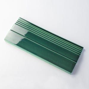 Może przedstawiać: Zielona płytka ceramiczna z błyszczącym wykończeniem i wypukłymi liniami. Płytka ma jasnozielony kolor u góry i ciemnozielony kolor u dołu.