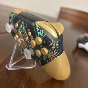 May include: A black video game controller featuring gold-colored buttons, triggers, and hand grips. The controller body is decorated with intricate green and gold graphic patterns and symbols. It is positioned on a clear plastic display stand.