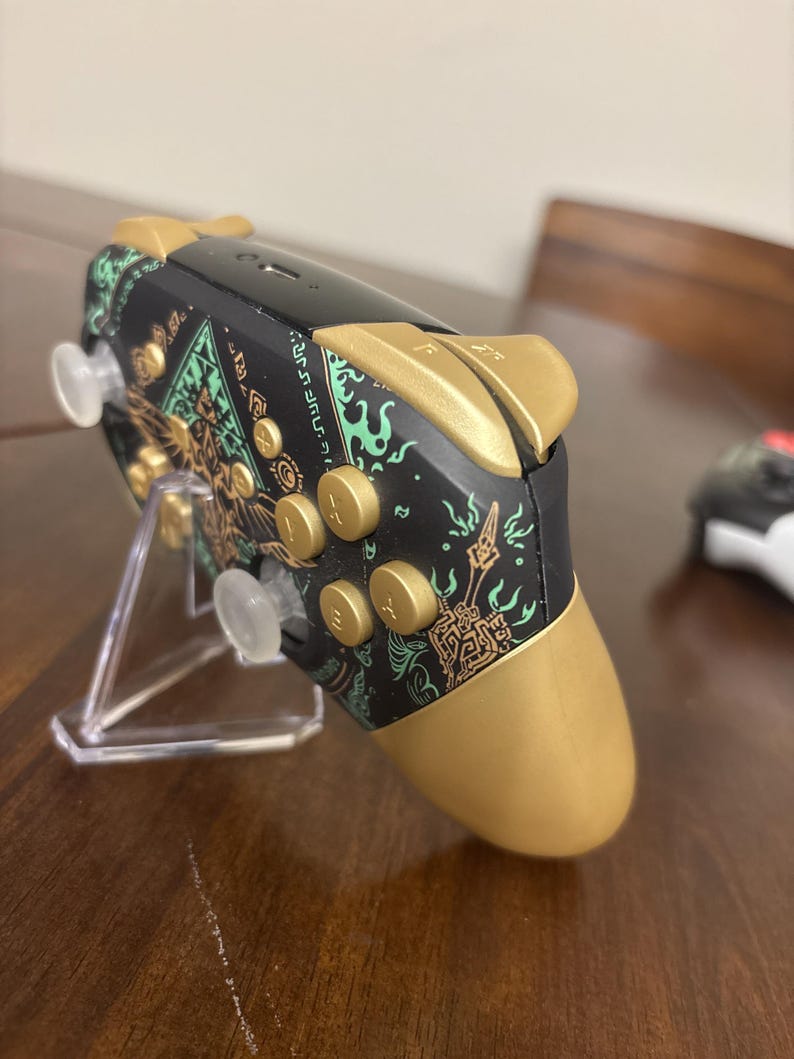 May include: A black video game controller featuring gold-colored buttons, triggers, and hand grips. The controller body is decorated with intricate green and gold graphic patterns and symbols. It is positioned on a clear plastic display stand.