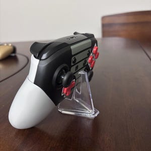 May include: A wireless video game controller featuring a black, white, and gray color scheme with bright red buttons and a directional pad. The controller is positioned on a clear, triangular plastic display stand, showing its side profile.