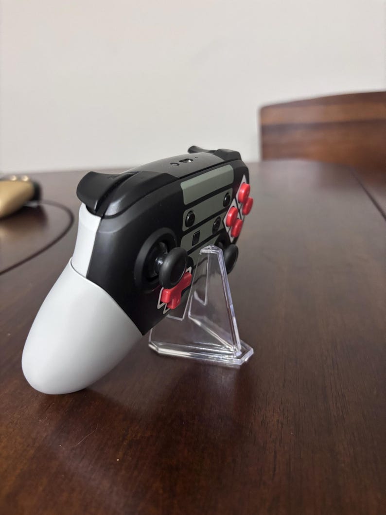 May include: A wireless video game controller featuring a black, white, and gray color scheme with bright red buttons and a directional pad. The controller is positioned on a clear, triangular plastic display stand, showing its side profile.