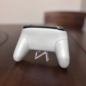 May include: A white wireless video game controller featuring a smooth, matte finish on the main body and two black shoulder buttons located at the top. The controller is positioned horizontally, showcasing its ergonomic grip handles and curved design.