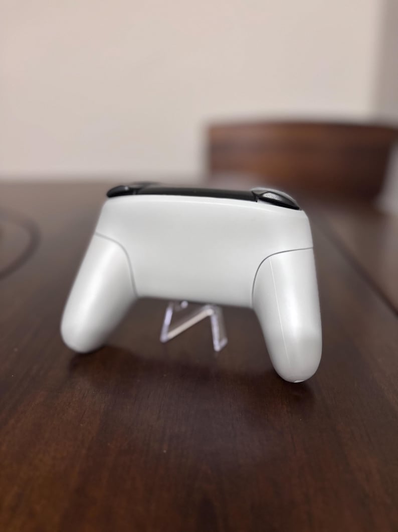 May include: A white wireless video game controller featuring a smooth, matte finish on the main body and two black shoulder buttons located at the top. The controller is positioned horizontally, showcasing its ergonomic grip handles and curved design.