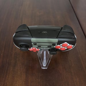 May include: A wireless video game controller featuring a black and olive green body with bright red action buttons and a directional pad. The top edge displays a central USB-C charging port and two black shoulder buttons on either side of the device.