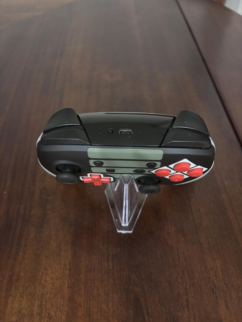 May include: A wireless video game controller featuring a black and olive green body with bright red action buttons and a directional pad. The top edge displays a central USB-C charging port and two black shoulder buttons on either side of the device.