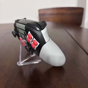 May include: A video game controller featuring a black and light gray body with bright red circular buttons. The controller is positioned on a clear plastic stand, showcasing its side profile, including the black shoulder triggers and analog sticks.