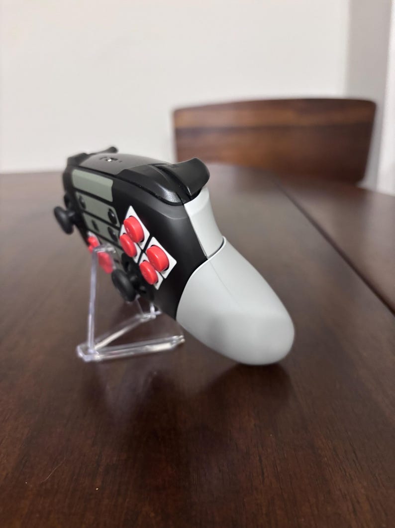 May include: A video game controller featuring a black and light gray body with bright red circular buttons. The controller is positioned on a clear plastic stand, showcasing its side profile, including the black shoulder triggers and analog sticks.