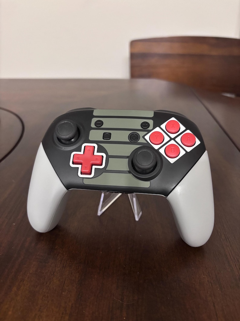 May include: A wireless video game controller featuring a black and light gray body with a central olive green panel. It includes a red directional pad, two black analog sticks, and four red action buttons labeled A, B, X, and Y on a white base.