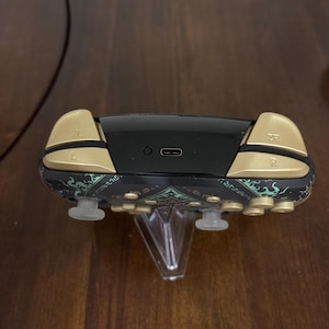 May include: A wireless game controller featuring a black body with teal and gold patterned accents. The top edge displays gold L, R, ZL, and ZR shoulder buttons, a central USB-C charging port, and a small circular sync button on a black panel.