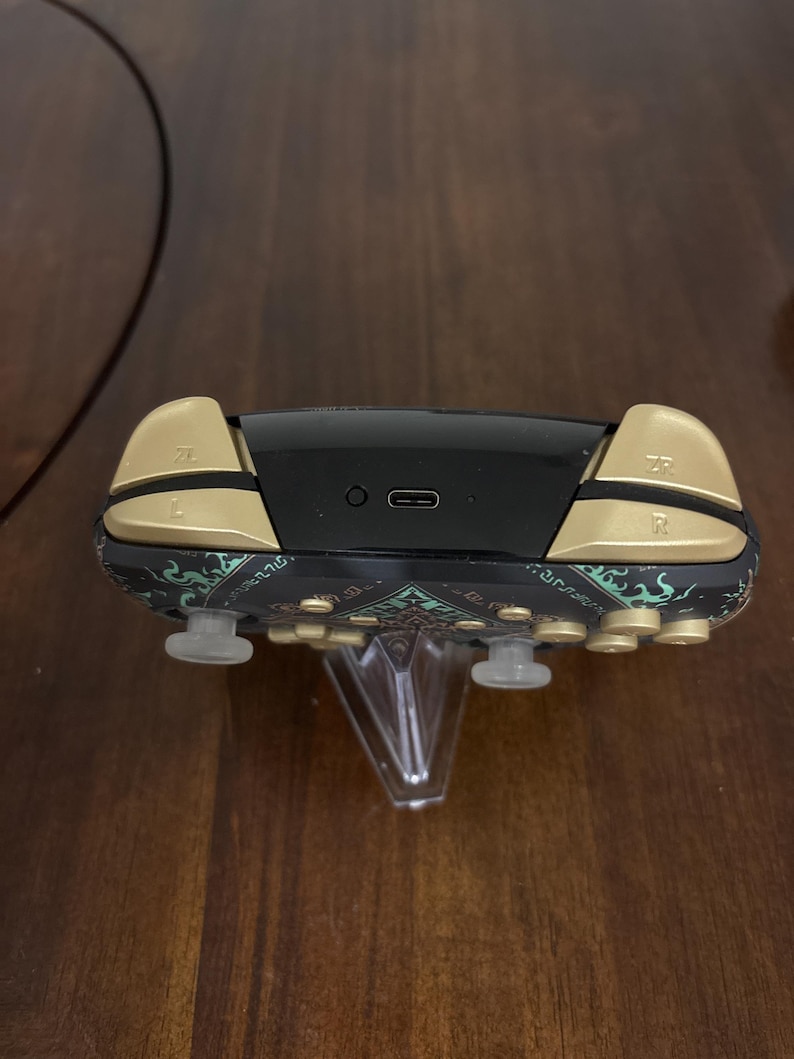 May include: A wireless game controller featuring a black body with teal and gold patterned accents. The top edge displays gold L, R, ZL, and ZR shoulder buttons, a central USB-C charging port, and a small circular sync button on a black panel.