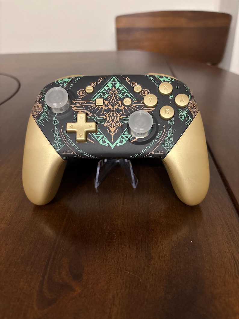May include: A wireless video game controller featuring a matte black faceplate with intricate gold and teal tribal-style patterns. The controller has gold-colored handles, a gold directional pad, gold action buttons labeled A, B, X, Y, and two clear joysticks.
