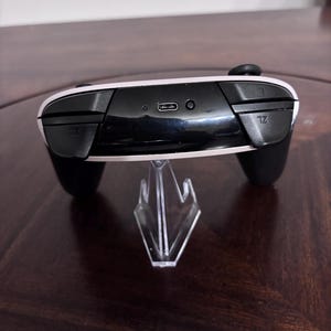 Pode incluir: Um controle de videogame preto e branco com uma porta de carregamento USB-C na parte superior, um pequeno bot&atilde;o circular de sincroniza&ccedil;&atilde;o e dois bot&otilde;es de ombro rotulados como L e ZL. O controle est&aacute; em um suporte de pl&aacute;stico transparente.