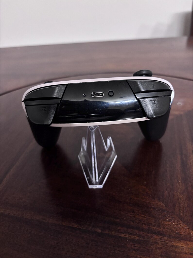 Pode incluir: Um controle de videogame preto e branco com uma porta de carregamento USB-C na parte superior, um pequeno bot&atilde;o circular de sincroniza&ccedil;&atilde;o e dois bot&otilde;es de ombro rotulados como L e ZL. O controle est&aacute; em um suporte de pl&aacute;stico transparente.